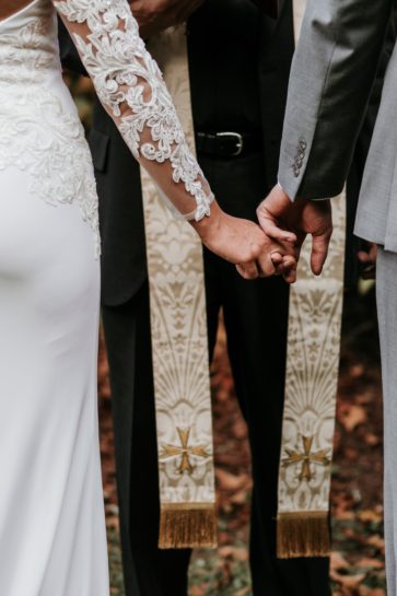 Para Młoda trzymająca się za ręce podczas ceremonii zaślubin w plenerze z księdzem