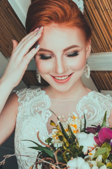 Uśmiechnięta ruda Panna Młoda prezentująca swój ślubny makijaż, trzymająca bukiet kwiatów