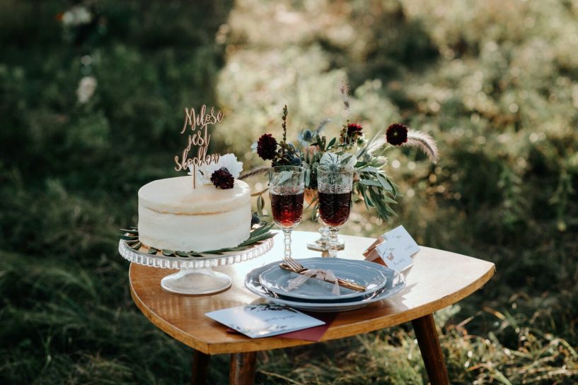 Stół z tortem ślubnym, zastawą, zaproszeniem, winietkami, kieliszkami z winem oraz bukietem w odcieniach bordo i zieleni