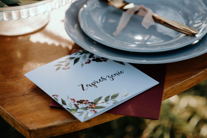 Zaproszenie ślubne z bordową kopertą przy zastawie stołowej ze złotymi sztućcami