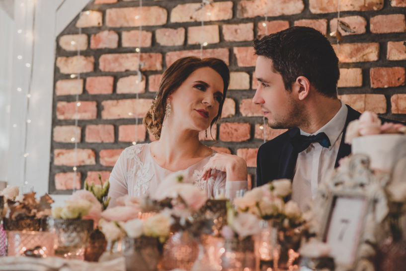 Elegancka Para Młoda, wpatrzona w siebie, siedząca przy stole bogato przyozdobionym kwiatami w wazonach i zapalonymi lampkami