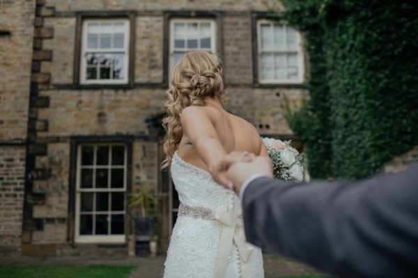 Ujęcie na tył fryzury ślubnej Panny Młodej trzymanej za rękę przez swojego wybranka